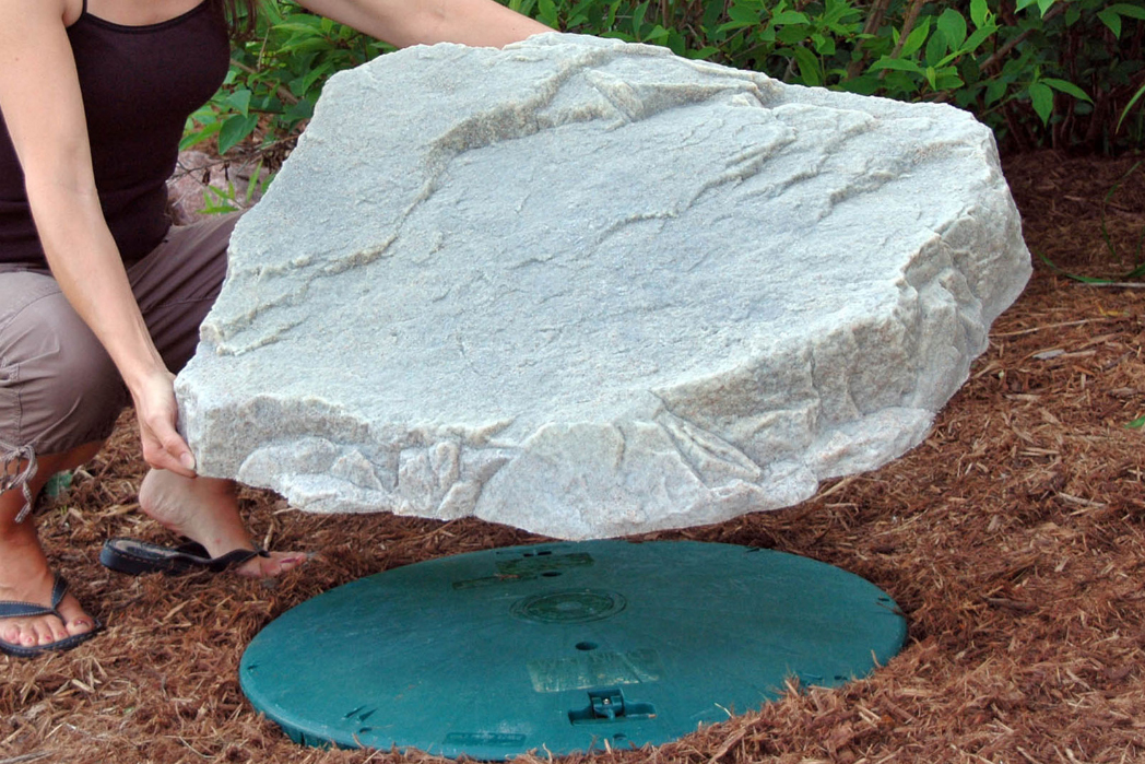 a photo of someone lifting faux rock model 108 up from a sewage cap.